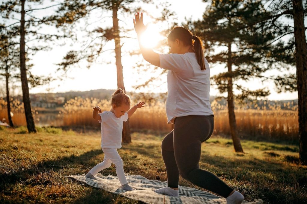 A woman outside playing with her child and practicing a yoga pose on grassland with sun in background